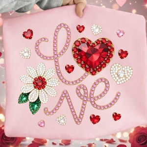Peut inclure: Un coussin rose avec le mot "LOVE" écrit en pierres précieuses roses. Le "O" est un grand cœur rouge, et il y a des motifs de cœurs et de fleurs plus petits. Le coussin est tenu par une personne.