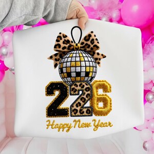Peut inclure: Un coussin blanc avec un motif du Nouvel An. Le motif comprend une boule disco, un nœud léopard et les chiffres "226" en noir, imprimé léopard et or. Les mots "Happy New Year" sont brodés en or.