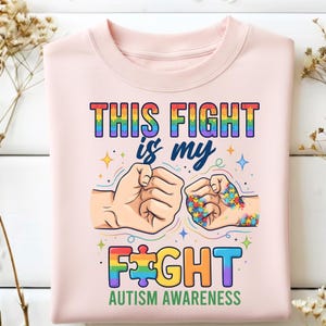 Puede incluir: Sudadera rosa claro con el texto "THIS FIGHT is my FIGHT" en colores arcoíris. El diseño incluye dos puños, uno con un patrón de rompecabezas, y las palabras "AUTISM AWARENESS".