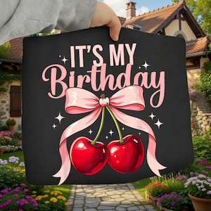 May include: Black graphic with the text "IT'S MY Birthday" in pink script. The design features two red cherries tied with a pink bow and ribbons, with star accents.