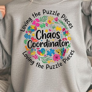 May include: A light gray sweatshirt with a colorful graphic that reads "Chaos Coordinator" surrounded by flowers, puzzle pieces, and other playful illustrations. The text "Loving the Puzzle Pieces" is arched around the design.