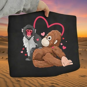 Op de afbeelding: Zwarte draagtas met een cartoonillustratie van twee apen, een roze hart en kleine roze hartjes. Eén aap is bruin, de andere is zwart-wit met een rood gezicht.