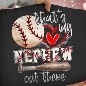 May include: Black t-shirt with a baseball graphic. The design includes a baseball, a red heart, and the words "That's my Nephew out there" in white. A wooden baseball bat is also featured.