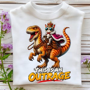 Peut inclure: Un sweat-shirt blanc à col rond orné d'un motif représentant un chat tigré portant une couronne, des lunettes et une cape rouge, chevauchant un dinosaure rayé orange et marron. Le texte sous le dinosaure indique "THIS IS AN OUTRAGE".