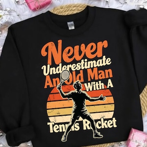 Peut inclure: Sweat-shirt noir avec le texte "Never Underestimate An Old Man With A Tennis Rocket" en orange et blanc. Une silhouette de joueur de tennis est présentée devant un motif de coucher de soleil.