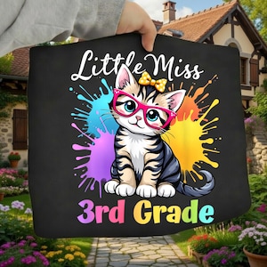 Puede incluir: Un artículo de tela negra con un gráfico de un gatito atigrado que lleva gafas rosas brillantes y un lazo amarillo con lunares. El texto Little Miss 3rd Grade aparece en letras coloridas, rodeado de salpicaduras de pintura en azul, morado y amarillo.