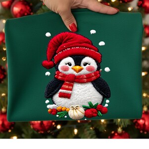 Peut inclure: Un sweat-shirt vert avec un pingouin brodé portant un bonnet et une écharpe rouges. Le pingouin a un ventre blanc, des ailes noires et un bec jaune. Des flocons de neige blancs et des baies festives entourent le pingouin, créant un thème de vacances.