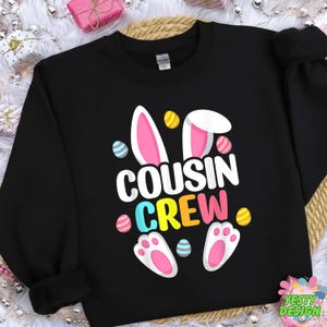 Peut inclure: Sweat-shirt noir avec l'inscription "COUSIN CREW" en blanc, jaune, rose et bleu. Le motif comprend des oreilles de lapin, des œufs de Pâques et des empreintes de pattes. Vêtement festif pour Pâques.