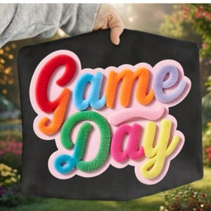 Peut inclure: Une pancarte noire avec l'inscription "Game Day" en lettres colorées et texturées. Les lettres sont rouges, orange, bleues, vertes, roses et jaunes, avec un contour rose. La pancarte est tenue par une personne.