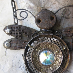 May include: A steampunk dragonfly pendant necklace, crafted from recycled metal components. The dragonfly design incorporates gears, bolts, and a central iridescent crystal. The pendant displays the words "KNOW YOUR WORTH".
