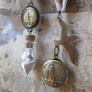 Puede incluir: Pendientes de temática costera con una pequeña botella de vidrio con arena, una concha y un dije de gaviota de metal. Los pendientes también incluyen un botón redondo dorado con un diseño de faro.