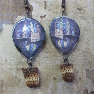 Può includere: Un paio di orecchini pendenti con design a mongolfiera e una cupola con motivi viola e blu. Ogni pallone è appeso a un gancio in metallo scuro e presenta un piccolo cestino all'uncinetto nei toni del marrone e del beige.