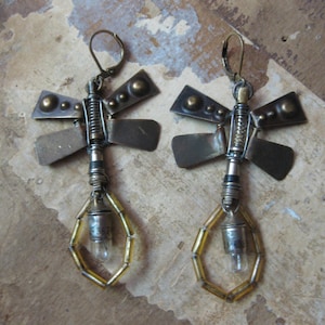 Könnte beinhalten: Ein Paar Ohrringe im Steampunk-Stil. Jeder Ohrring hat ein Libellen-Design mit Metallflügeln und einer kleinen Glühbirne in einem geometrischen Rahmen. Die Ohrringe haben ein Bronze- und Silberschema.