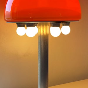 May include: A table lamp featuring a large, vibrant orange dome-shaped glass shade. The lamp has a central brushed metal cylindrical stem supported by a circular white marble base. Four small, round, glowing white light bulbs are visible underneath.