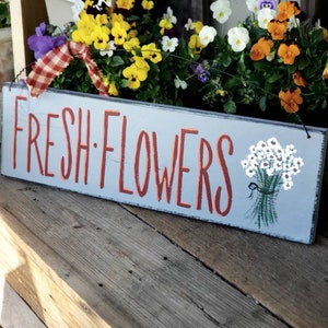 May include: A rectangular wooden sign with the words "FRESH FLOWERS" in red lettering. A painted bouquet of white flowers with green stems and a black ribbon is on the right. The sign is light blue.