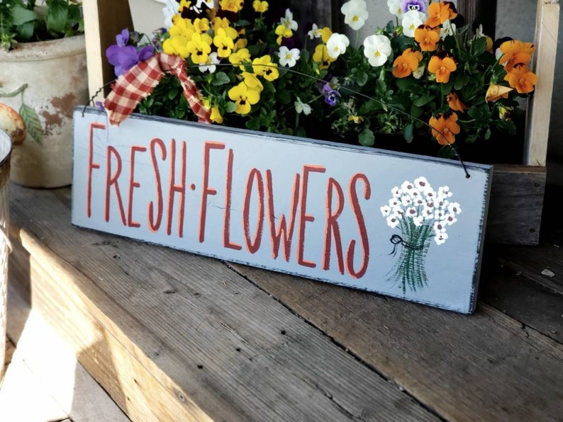 May include: A rectangular wooden sign with the words "FRESH FLOWERS" in red lettering. A painted bouquet of white flowers with green stems and a black ribbon is on the right. The sign is light blue.