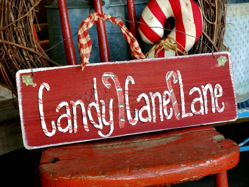 May include: A red wooden sign with the words "Candy Cane Lane" in white script. The letters are designed to look like candy canes. The sign is decorated with glitter and a red and white checkered ribbon.
