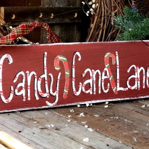 May include: A rectangular, rustic wooden sign painted red with the words "Candy Cane Lane" in white script. Candy cane illustrations are incorporated into the lettering. A plaid ribbon is attached at the top for hanging. The sign is a festive holiday decoration.