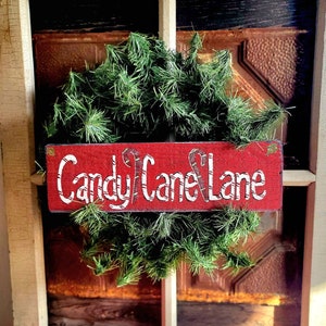 May include: A Christmas wreath with a red sign that reads "Candy Cane Lane" in white lettering. The sign has candy cane illustrations. The wreath is made of green pine needles and is set in a rustic window frame.