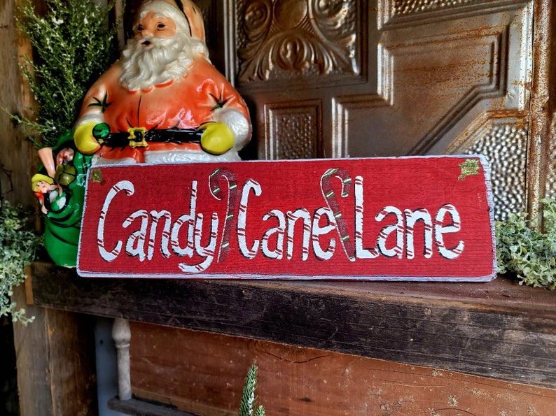 May include: A rectangular red wooden sign with the words "Candy Cane Lane" in white script. The letters are decorated with candy cane stripes. A Santa Claus figurine and a green sack are in the background.