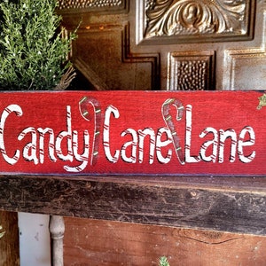 May include: A rectangular, red wooden sign with the words "Candy Cane Lane" in white, featuring candy cane accents. The sign is decorated with small green accents and is placed on a rustic wooden shelf, evoking a festive holiday theme.