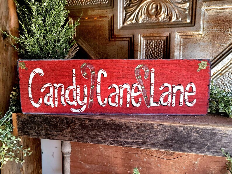 May include: A rectangular, red wooden sign with the words "Candy Cane Lane" in white, featuring candy cane accents. The sign is decorated with small green accents and is placed on a rustic wooden shelf, evoking a festive holiday theme.