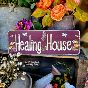 May include: A rectangular wooden sign with the words "Healing House" in white script. The sign is eggplant-colored with painted flowers, bees, and butterflies. The sign is surrounded by flowers and vintage metal containers.