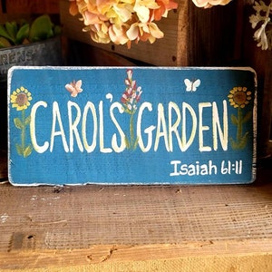 May include: A rectangular, hand-painted wooden sign with a distressed blue background. The sign reads "Carol's Garden" in white script, with sunflowers and butterflies. The text "Isaiah 61:11" is also present. The sign is a decorative garden accent.