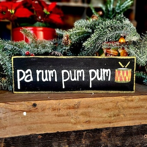 May include: A rectangular black wooden sign with gold trim features the phrase "pa rum pum pum" in white lettering, accompanied by a drum illustration. The sign is set against a backdrop of Christmas greenery and red poinsettias.