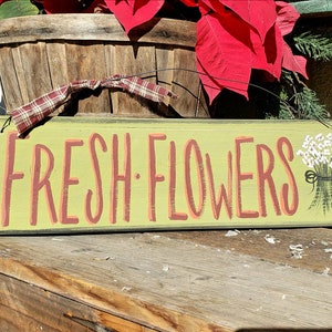 May include: A rectangular wooden sign with the words "FRESH FLOWERS" in coral-colored letters on a light green background. A small bouquet of white flowers is painted on the right side. The sign is placed on a wooden surface.