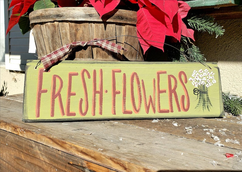 May include: A rectangular wooden sign with the words "FRESH FLOWERS" in coral-colored letters on a light green background. A small bouquet of white flowers is painted on the right side. The sign is placed on a wooden surface.