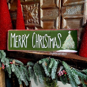 May include: A green wooden sign with the words "MERRY CHRISTMAS" in white lettering and a painted Christmas tree. The sign is decorated with a garland of pine needles, berries, and pine cones, and red glittery cone-shaped trees.