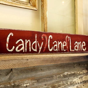 May include: A rustic, rectangular wooden sign with the words "Candy Cane Lane" in white, candy cane-striped lettering. The sign is painted a deep red and decorated with holly and berries, evoking a festive holiday theme.