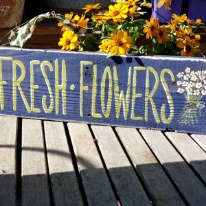 May include: A rectangular wooden sign painted navy blue with the words "FRESH FLOWERS" in light green lettering. A bouquet of white flowers is painted on the right side. The sign is hanging with a green and white checkered ribbon.