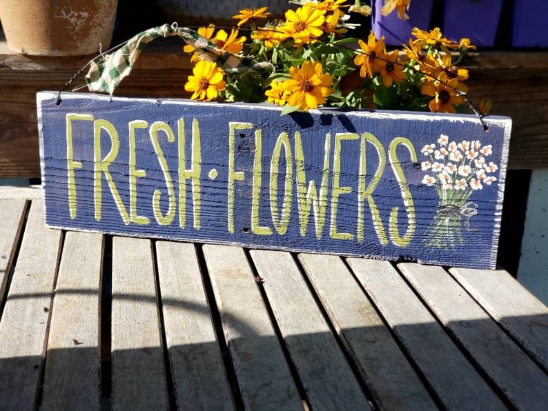 May include: A rectangular wooden sign painted navy blue with the words "FRESH FLOWERS" in light green lettering. A bouquet of white flowers is painted on the right side. The sign is hanging with a green and white checkered ribbon.