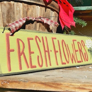 May include: A rectangular wooden sign with the words "FRESH FLOWERS" in red letters on a light green background. A small bouquet of white flowers is painted on the right side. The sign is decorated with a red bow and plaid ribbon.