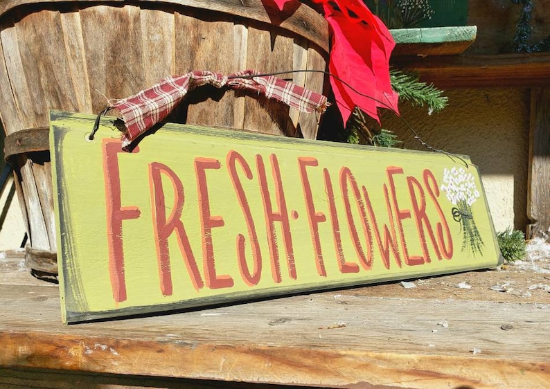 May include: A rectangular wooden sign with the words "FRESH FLOWERS" in red letters on a light green background. A small bouquet of white flowers is painted on the right side. The sign is decorated with a red bow and plaid ribbon.