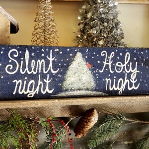 May include: A rectangular wooden sign with the words "Silent night Holy night" in white script. The sign features a painted Christmas tree and falling snow on a dark blue background. A small red bird is perched on the tree.