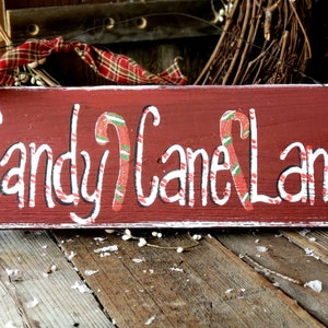 May include: A rectangular, rustic wooden sign painted a deep red with white lettering that reads "Candy Cane Lane." The sign features candy cane illustrations and is decorated with a plaid ribbon and gold glitter accents, evoking a festive Christmas theme.