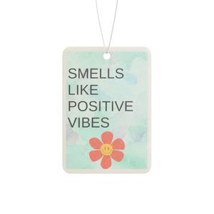 Può includere: Deodorante per ambienti rettangolare con sfondo acquerello nei toni del verde e del blu. Il testo "SMELLS LIKE POSITIVE VIBES" è stampato in nero. Un fiore rosso con una faccina sorridente gialla è in basso.
