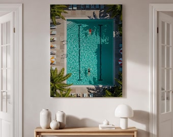 May include: A framed print showing an aerial view of a swimming pool with two swimmers. The turquoise water is divided by black lane lines. Palm trees and sun loungers surround the pool. One swimmer wears a red swimsuit, the other a black swimsuit.