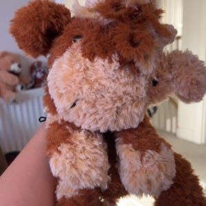 May include: A plush toy cow with brown and cream-colored fur. The stuffed animal has a fluffy appearance, with brown patches on its ears, around its eyes, and on its hooves. It has a small tail and a friendly expression.
