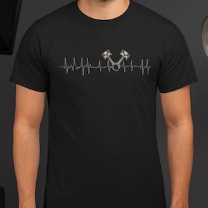Op de afbeelding: Zwart T-shirt met een zilveren hartslagontwerp en twee zuigers. De zuigers zijn realistisch gearceerd. Het ontwerp is gecentreerd op de borst van het shirt.