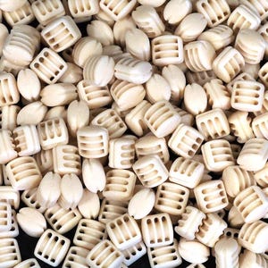May include: An assortment of off-white beads, comprising square and oval shapes. The square beads feature a ribbed design, and the oval beads are smooth. The beads are scattered, creating a textured appearance.