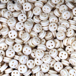 May include: A close-up of a collection of off-white, circular buttons, each with three holes. The buttons have a slightly raised, double-layered design. The buttons are scattered, filling the frame.