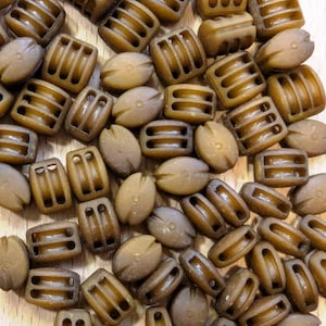 May include: A selection of small, brown beads in different forms. Some are barrel-shaped with three rectangular apertures, while others are oval with a small indentation. The beads have a matte finish and are scattered on a wooden surface.