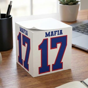Op de afbeelding: Een witte vierkante memoblokkubus met de tekst MAFIA en het getal 17 in vetgedrukt blauw met een rode omlijning op twee zichtbare zijden. De papieren kubus staat op een houten bureau naast een koffiemok en een notitieboek.