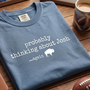Op de afbeelding: Een blauw T-shirt met de tekst "probably thinking about Josh...again" en een witte buffelafbeelding. Het shirt wordt gepresenteerd met een voetbal, een mok en een notitieboek.
