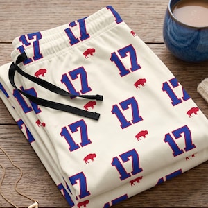 May include: Off-white lounge pants featuring a repeating pattern of the blue number 17 and small red buffalo silhouettes. The pants have a black drawstring at the elastic waistband. A gold-tone necklace with the number 17 is also visible.