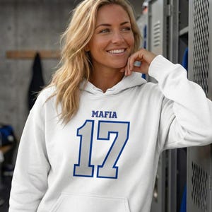 Puede incluir: Sudadera blanca con la palabra "MAFIA" encima del n&uacute;mero "17" en azul y gris. La sudadera tiene un bolsillo delantero y una capucha con cord&oacute;n. La persona de la imagen est&aacute; sonriendo.